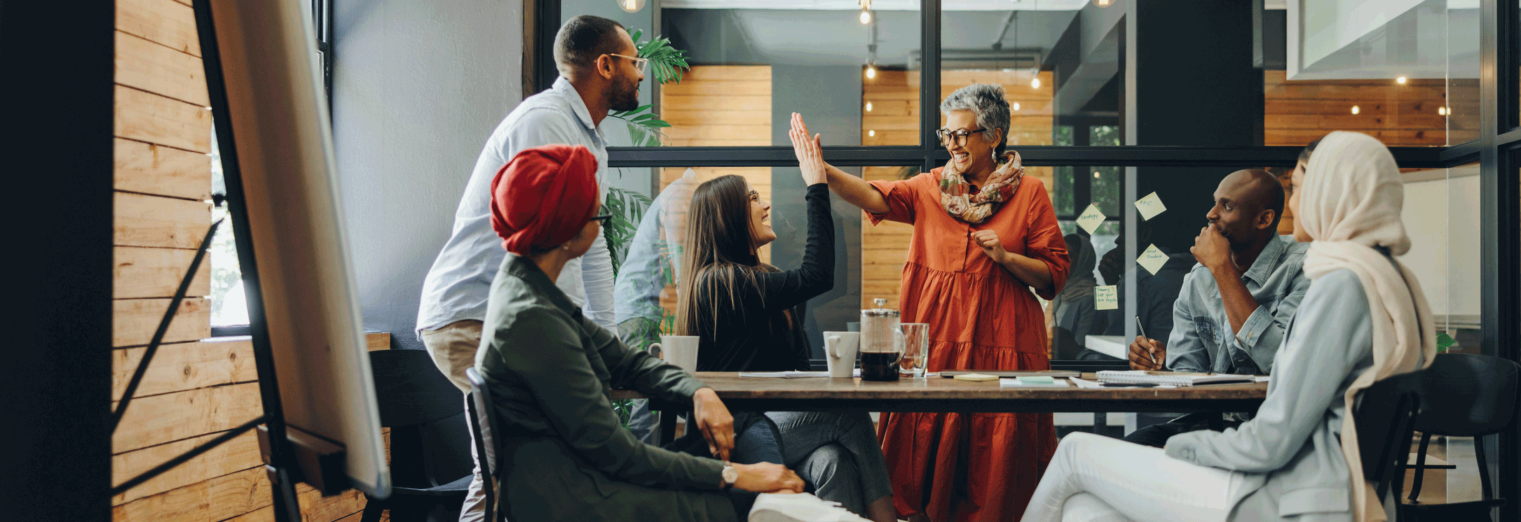 A group of 6 people engaged in a discussion in an office boardroom, with two women sharing a high-five.